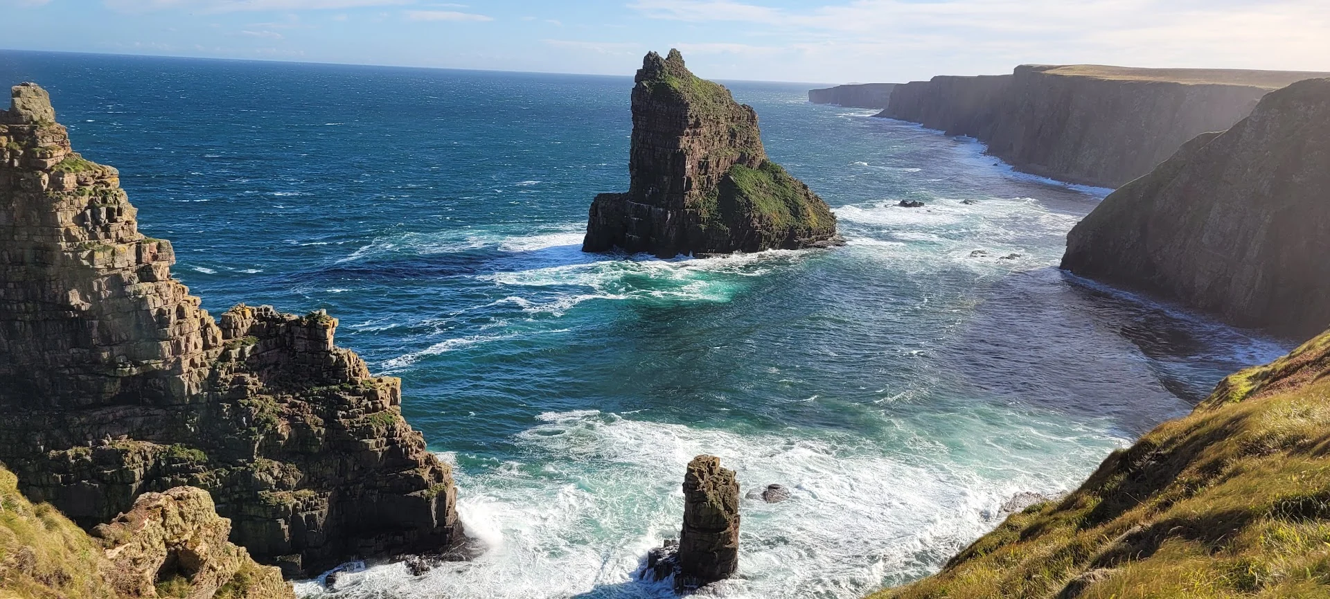 Dark sea stacks rise from blue-green water below a long line of sunlit cliffs.