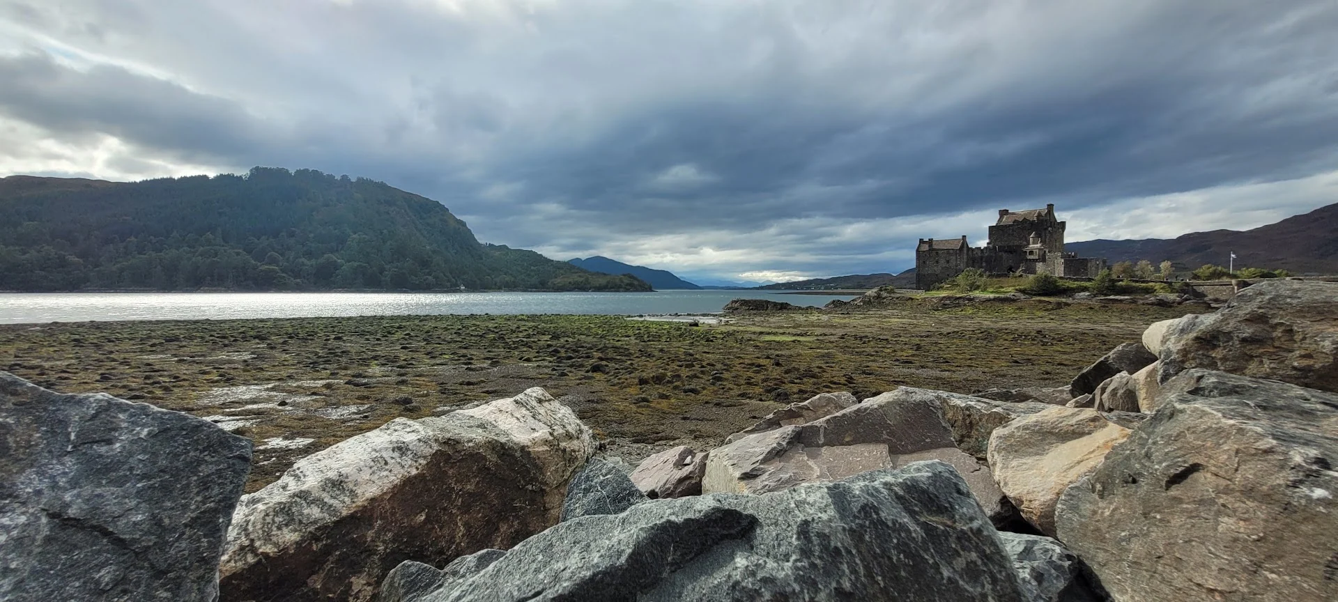 Eilean Donan Castle stands beyond a rocky tidal shore under heavy clouds in the Scottish Highlands.
